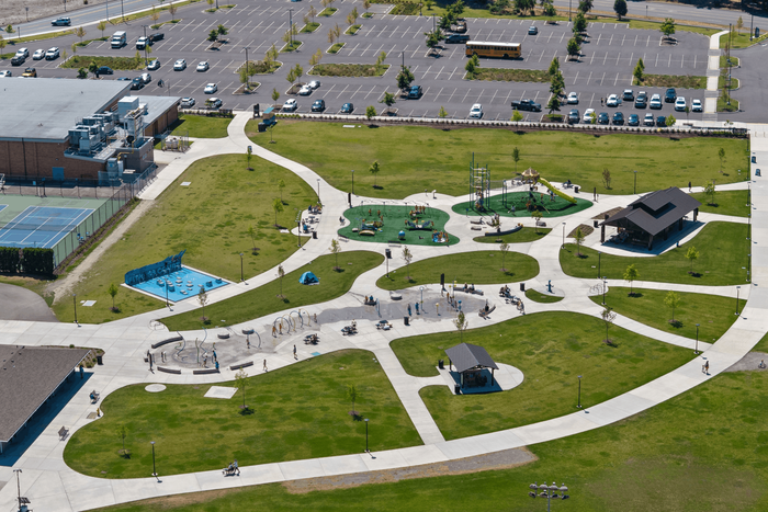 Aerial view of vibrant community park with winding paths, lush greenery, playgrounds, and sports courts surrounded by a large parking area.