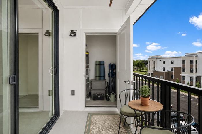 Charming balcony with a cozy seating area, vibrant potted plant, and open storage nook, overlooking a spacious, modern apartment complex under a bright blue sky.