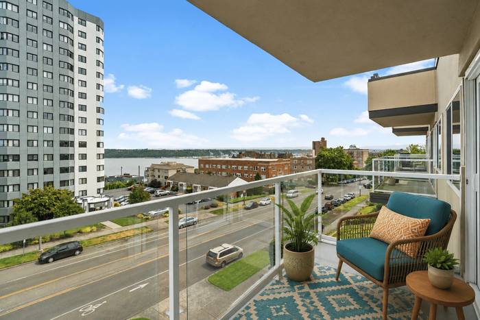 Charming balcony view from a modern apartment featuring cozy seating, vibrant potted plants, and a panoramic cityscape against a tranquil river backdrop.