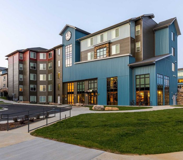 Modern multi-story apartment building with distinctive blue and brown exterior and large windows, set in a landscaped area with a vibrant atmosphere.