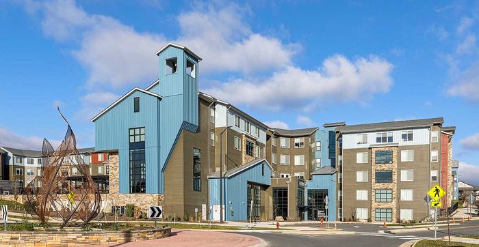 Modern apartment complex with unique architectural design, featuring blue and beige tones, large windows, and a distinctive sculpture in the foreground under a clear blue sky.