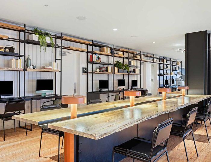 Modern communal workspace with sleek wooden tables, ambient lighting, and computers, framed by stylish shelving and decor.