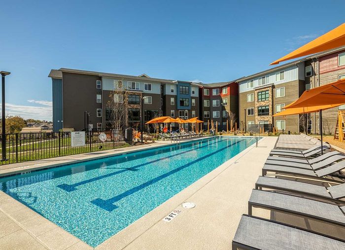 Luxurious apartment complex featuring a sparkling outdoor swimming pool with sun loungers and vibrant orange umbrellas, surrounded by modern architecture.
