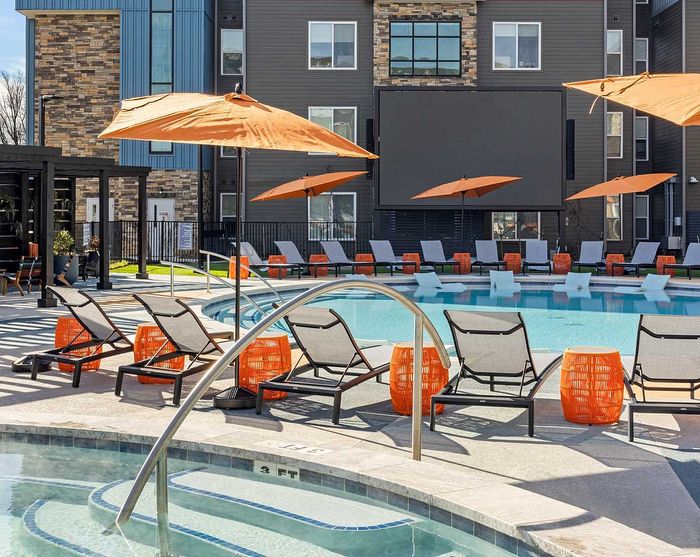 Luxurious apartment complex featuring a spacious outdoor pool area with stylish lounge chairs, vibrant orange umbrellas, and a large outdoor screen.