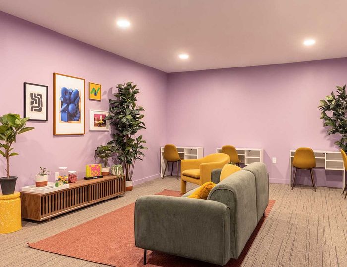 Vibrant living area with lavender walls, modern artwork, green sofa, and cozy yellow chairs. Ideal for relaxation and creativity.