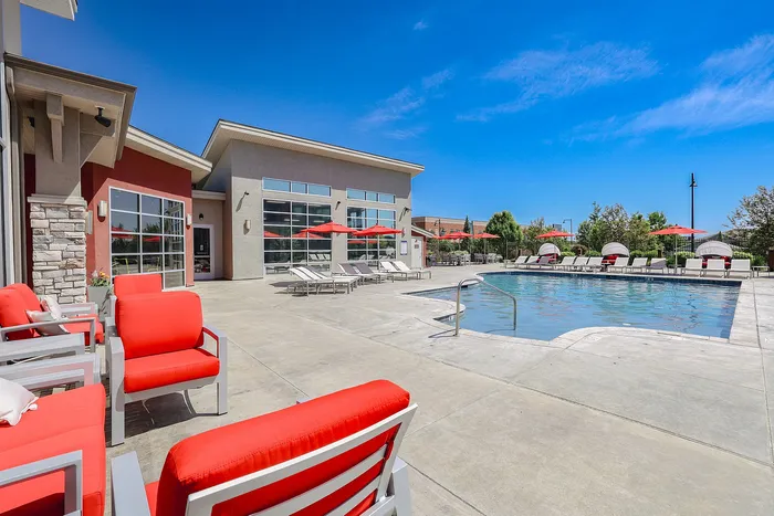 Luxurious outdoor pool area featuring modern red lounge chairs and umbrellas, set against a vibrant blue sky and contemporary clubhouse backdrop.