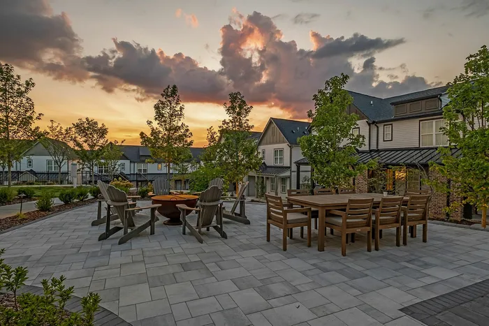 Charming outdoor patio with cozy fire pit and elegant dining set, framed by lush trees against a stunning sunset sky.