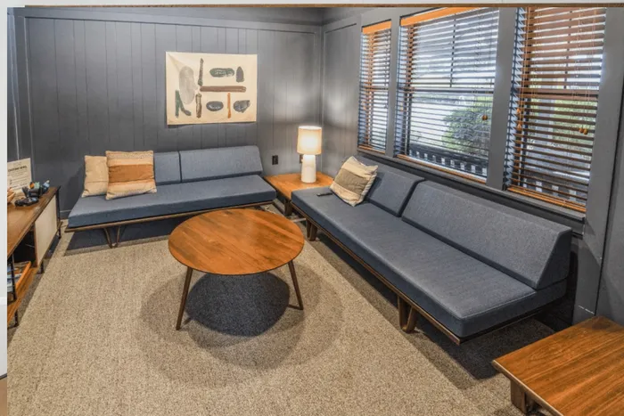 Inviting living room with modern blue sofas, elegant wooden coffee table, and stylish wall art, bathed in natural light from large windows with wooden blinds.