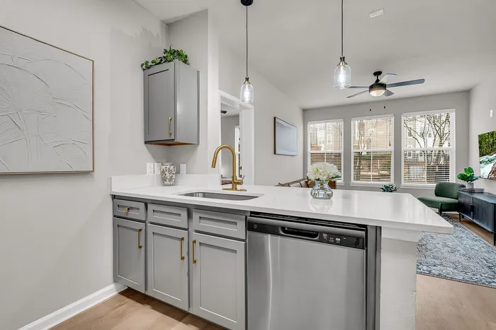 Elegant apartment interior showcasing a modern kitchen with sleek gray cabinets, a luxurious gold faucet, and stainless-steel appliances, opening to a bright living area with large windows.