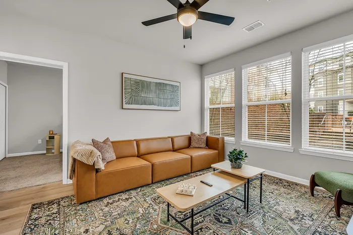 Bright and inviting living room with a plush leather sofa, modern ceiling fan, and ample natural light from large windows.