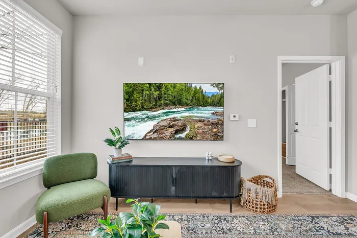 Modern living room with stylish green accent chair, elegant black media console, and a wall-mounted TV displaying a serene nature scene. Bright natural light streams through large windows, enhancing the cozy and inviting atmosphere.