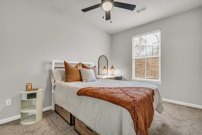 Cozy bedroom featuring a stylish bed with warm-toned pillows and a textured throw, complemented by a modern ceiling fan and soft natural light.