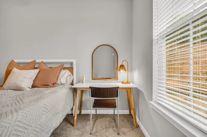 Cozy bedroom nook featuring a stylish workspace with a white desk, modern chair, and elegant lamp, complemented by plush pillows and natural light from the window.
