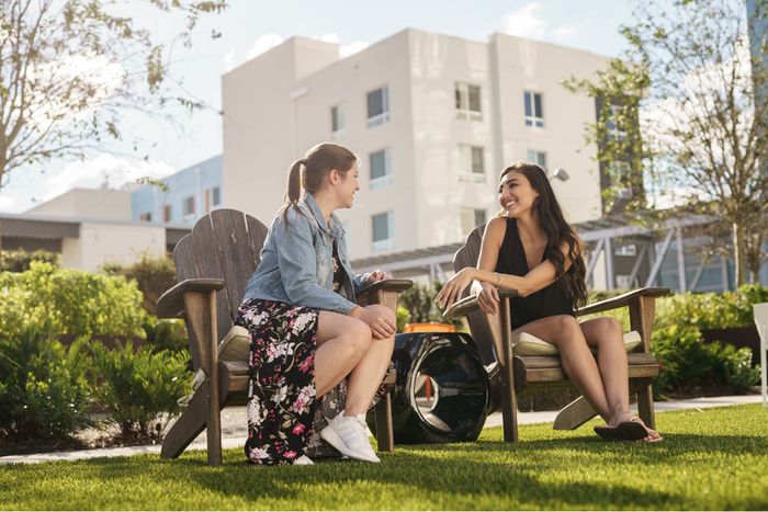 Bright outdoor scene with two people enjoying a friendly conversation on wooden chairs, framed by lush greenery and modern apartment buildings in the background.