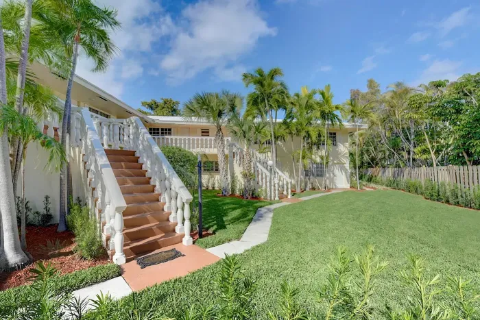Inviting apartment complex with a sunlit pool surrounded by lush tropical greenery and modern white buildings.