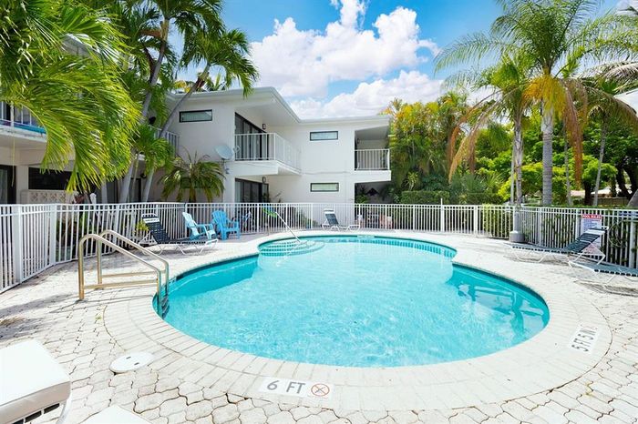 Inviting apartment complex with a sunlit pool surrounded by lush tropical greenery and modern white buildings.