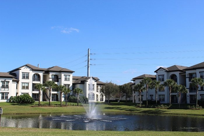 Elegant apartment complex surrounded by lush greenery, featuring a serene pond with a charming fountain under a clear blue sky.