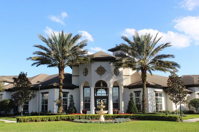 Elegant apartment complex entrance with palm trees and a decorative fountain, set against a bright blue sky.
