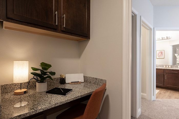 Cozy and modern workspace with a granite desk, warm lighting, and sleek cabinetry, adjacent to a well-lit hallway leading to a chic bathroom.