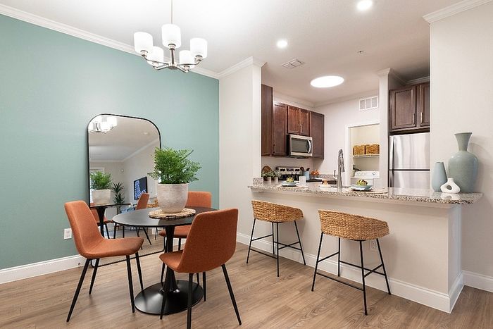 Modern dining and kitchen area featuring elegant wood flooring, stylish bar stools, orange chairs, and a chic round table with greenery.