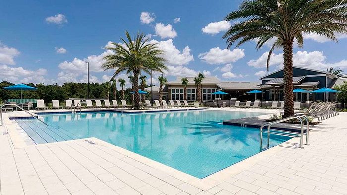 Luxurious resort-style pool area with palm trees, sun loungers, and a modern clubhouse under a vibrant blue sky.