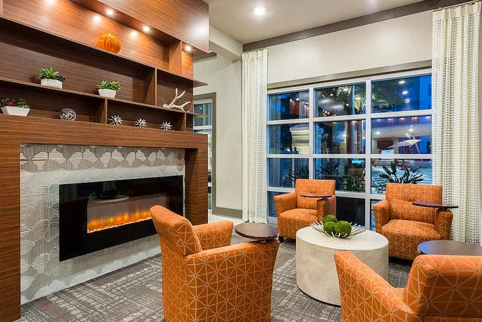 Chic and cozy living area with modern orange armchairs, elegant wooden shelves, and a stylish fireplace, enhanced by large windows offering natural light.