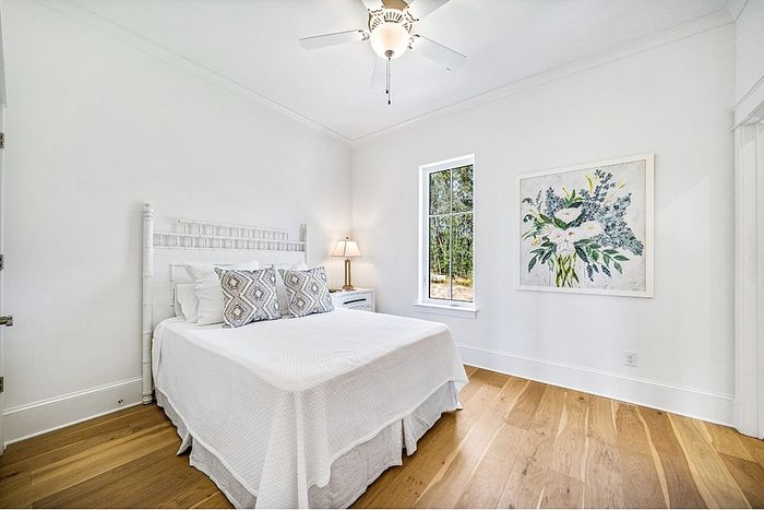 Bright, airy bedroom with a stylish white bed and elegant artwork, featuring hardwood floors and a large window overlooking greenery.