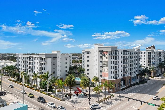 Modern apartment complex with elegant design, spacious balconies, and lush palm-lined entrance, set against a vibrant blue sky.