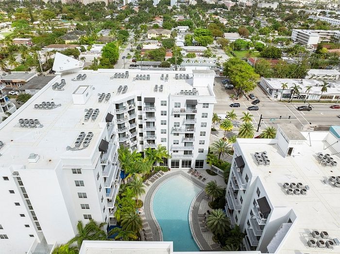 Aerial view of modern apartment complex featuring lush landscaping, sparkling central pool, and panoramic cityscape.