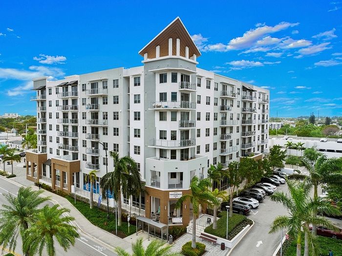 Modern apartment building with sleek design, spacious balconies, and lush palm trees under a vibrant blue sky.