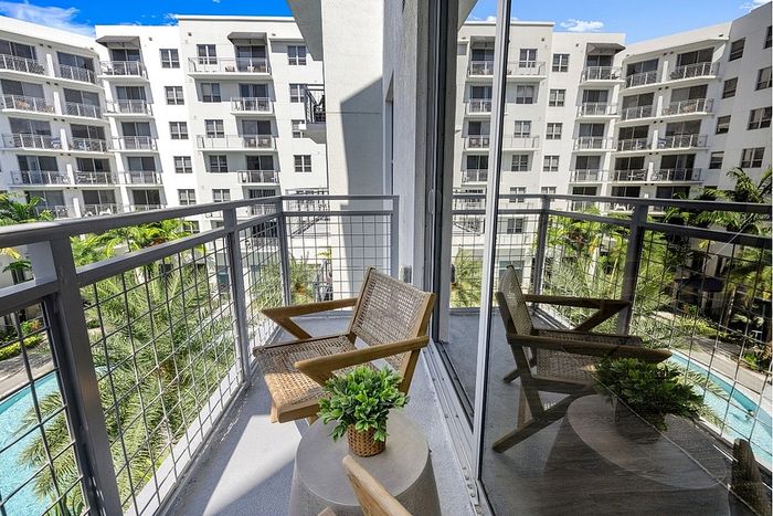 Chic apartment balcony with a cozy chair and lush greenery, offering stunning views of a modern complex and sparkling pool.