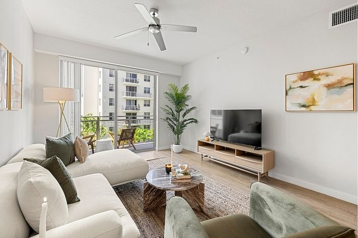 Bright, modern living room with stylish furnishings, large TV, cozy seating, and a spacious balcony overlooking lush greenery.