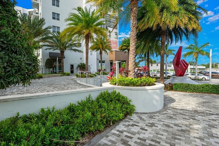 Contemporary apartment entrance surrounded by lush palm trees and modern art sculpture, offering a vibrant lifestyle.