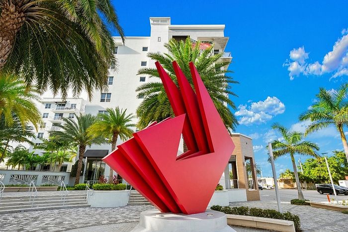 Modern apartment exterior featuring a striking red sculpture, surrounded by lush palm trees, under a clear blue sky.