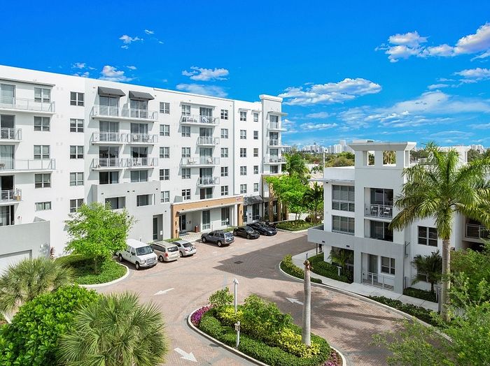 Modern apartment complex with lush landscaping, featuring a white exterior and large windows under a vibrant blue sky.
