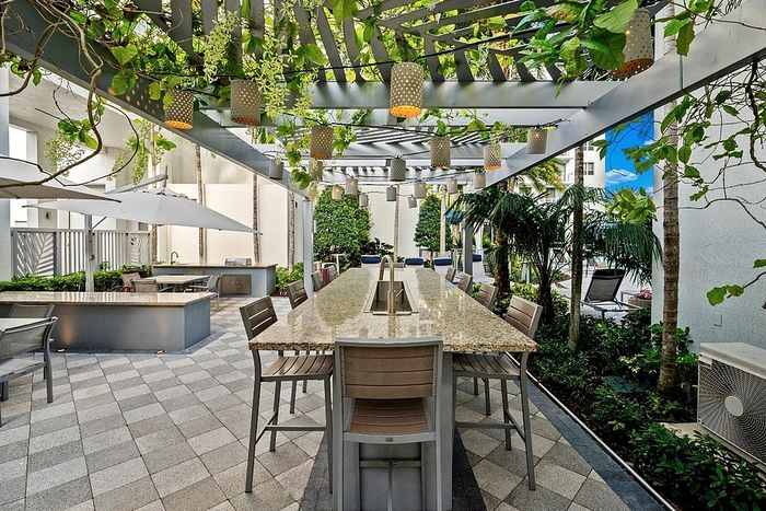 Elegant outdoor terrace featuring a long granite countertop, modern bar stools, lush greenery, and stylish pergola with hanging lights.