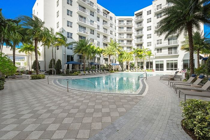 Elegant apartment complex featuring a pristine pool, lush palm trees, and modern architecture under a bright blue sky.