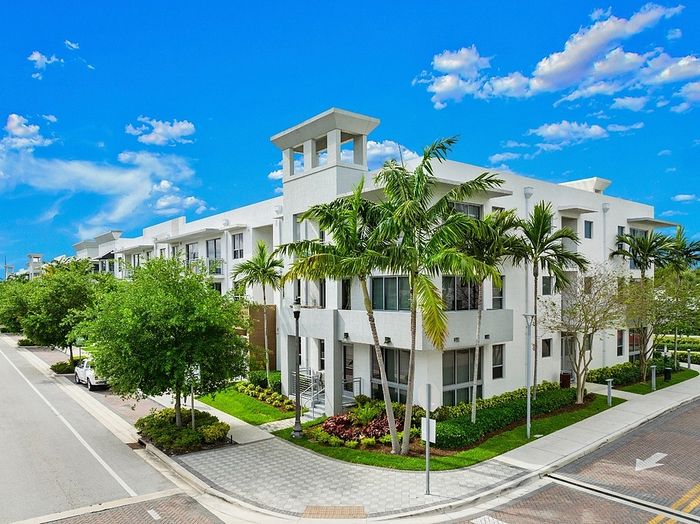 Modern apartment building in a tropical setting, featuring lush palm trees and pristine landscaping under a vibrant blue sky.