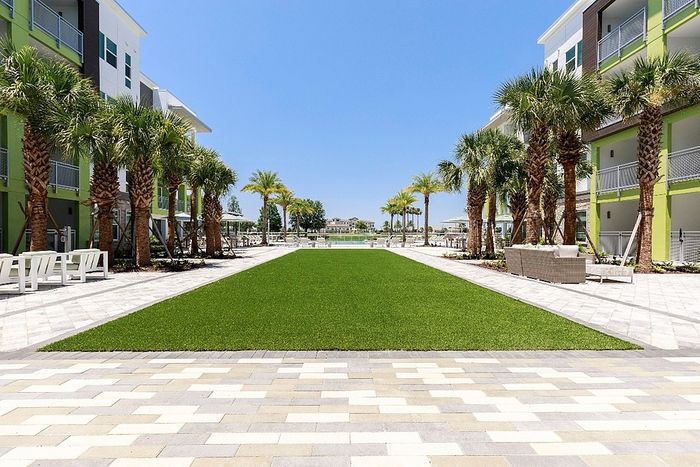 Luxurious outdoor courtyard with lush green lawn, lined with tall palm trees, surrounded by modern apartment buildings under a clear blue sky.