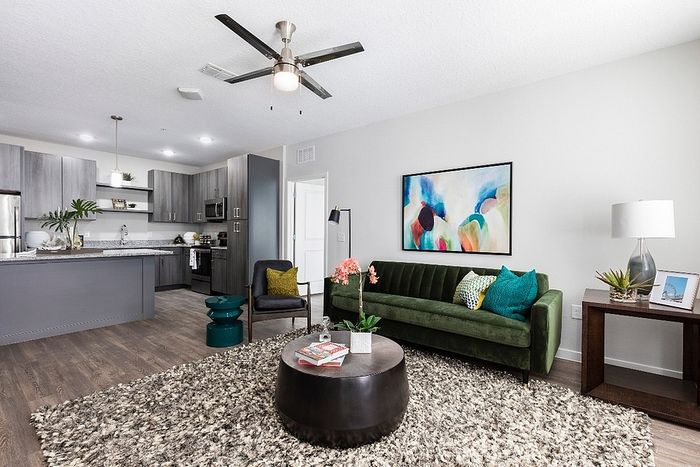 Modern open-plan living room with sleek kitchen, featuring a plush green sofa, abstract art, and cozy decor on elegant wood flooring.