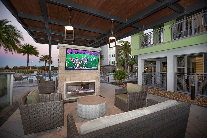 Chic outdoor lounge area featuring cozy wicker seating, a flat-screen TV, and a fireplace, perfect for relaxing by the pool with palm tree views.
