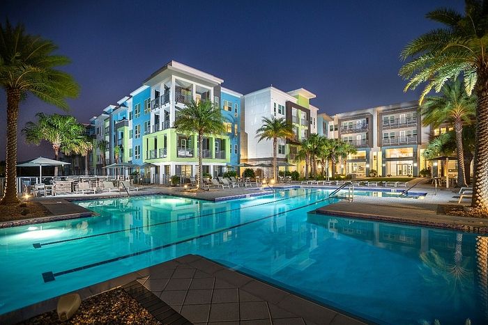 Luxurious apartment complex with vibrant modern design, featuring a stunning illuminated pool and lush palm trees at dusk.