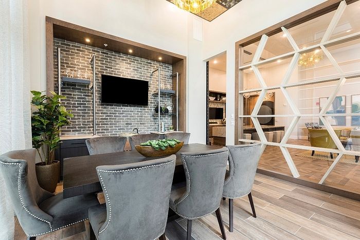 Chic dining area with elegant gray chairs, stylish brick accent wall, and modern decor, complemented by a view into a cozy, inviting living space.