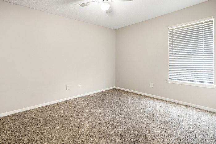 Bright, carpeted bedroom with natural light streaming through the window, featuring neutral tones and a ceiling fan.