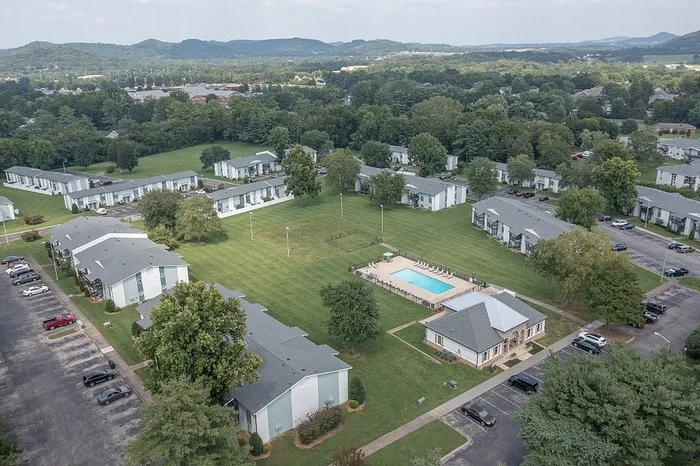 Scenic aerial view of a welcoming apartment complex with lush green spaces, a central swimming pool, and multiple modern apartment buildings surrounded by trees and hills.