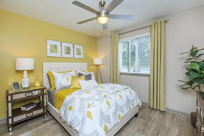 Bright and inviting bedroom with a modern yellow accent wall, stylish bedding, and elegant decor. Features a ceiling fan, large window for natural light, and sleek wooden flooring. Perfect blend of comfort and sophistication.