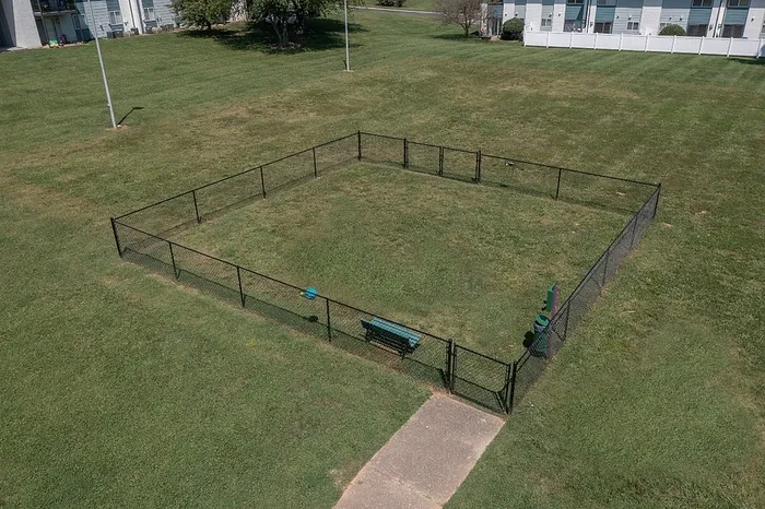 Charming green outdoor space featuring a fenced dog park area with a bench, surrounded by lush lawns and nearby residences.