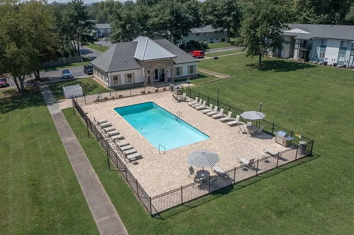 Aerial view of a luxurious apartment complex featuring a sparkling swimming pool, elegant poolside seating, and a charming clubhouse surrounded by lush green lawns.