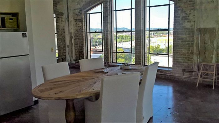 Chic industrial loft dining area with exposed brick walls, large windows offering scenic views, and a stylish wooden table with white chairs.
