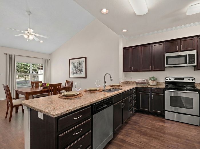 Elegant open kitchen with granite countertops and stainless steel appliances, adjacent to a sunlit dining area with wooden furniture.